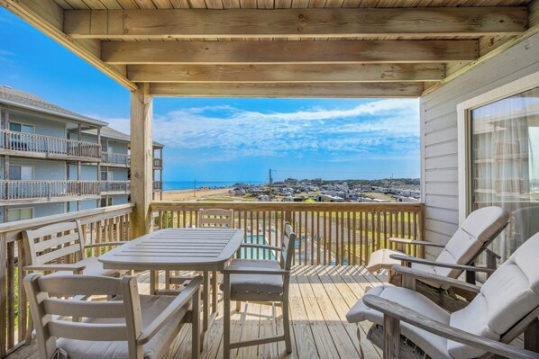 A wooden deck with a table and chairs, overlooking a beach and a parking area.