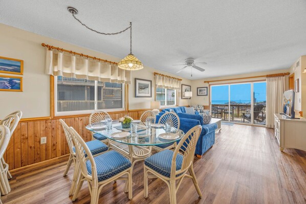 A dining area with a glass table and wicker chairs, a blue sofa, and a view of the ocean through large windows. - Semi-Oceanfront Condo w/ Private Balcony, Resort Pool, Elevator, Beach Boardwalk (Rodanthe)