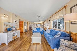 Un salon spacieux avec un grand canapé bleu, une table basse blanche et un plancher en bois. Au fond, on aperçoit une cuisine donnant sur un bâtiment à l'extérieur.
