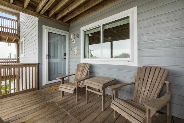 A wooden deck with two Adirondack chairs and a small table, a door, and a window with a view.
