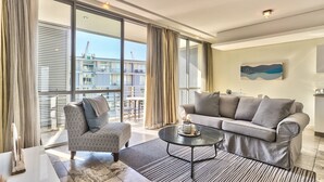 Een moderne woonkamer met een grijze bank, een fauteuil met dessin, en een salontafel. Grote ramen bieden uitzicht op de stad.