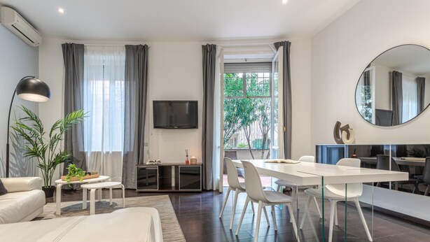 Ein modernes Wohnzimmer mit einem weißen Sofa, einem Esstisch mit Stühlen, einem an der Wand montierten Fernseher und einem großen Fenster mit Vorhängen.