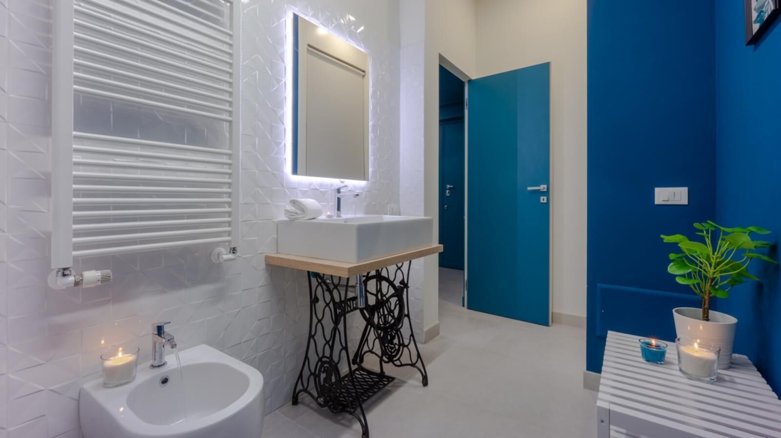 Een moderne badkamer met een witte wasbak, spiegel en toiletbril. Achtergrond is een blauwe deur en rechts staat een potplant.