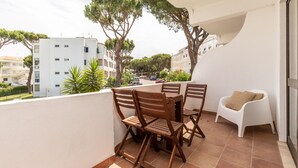 Un balcón con una mesa y sillas de madera, una silla blanca y vistas a edificios y árboles.