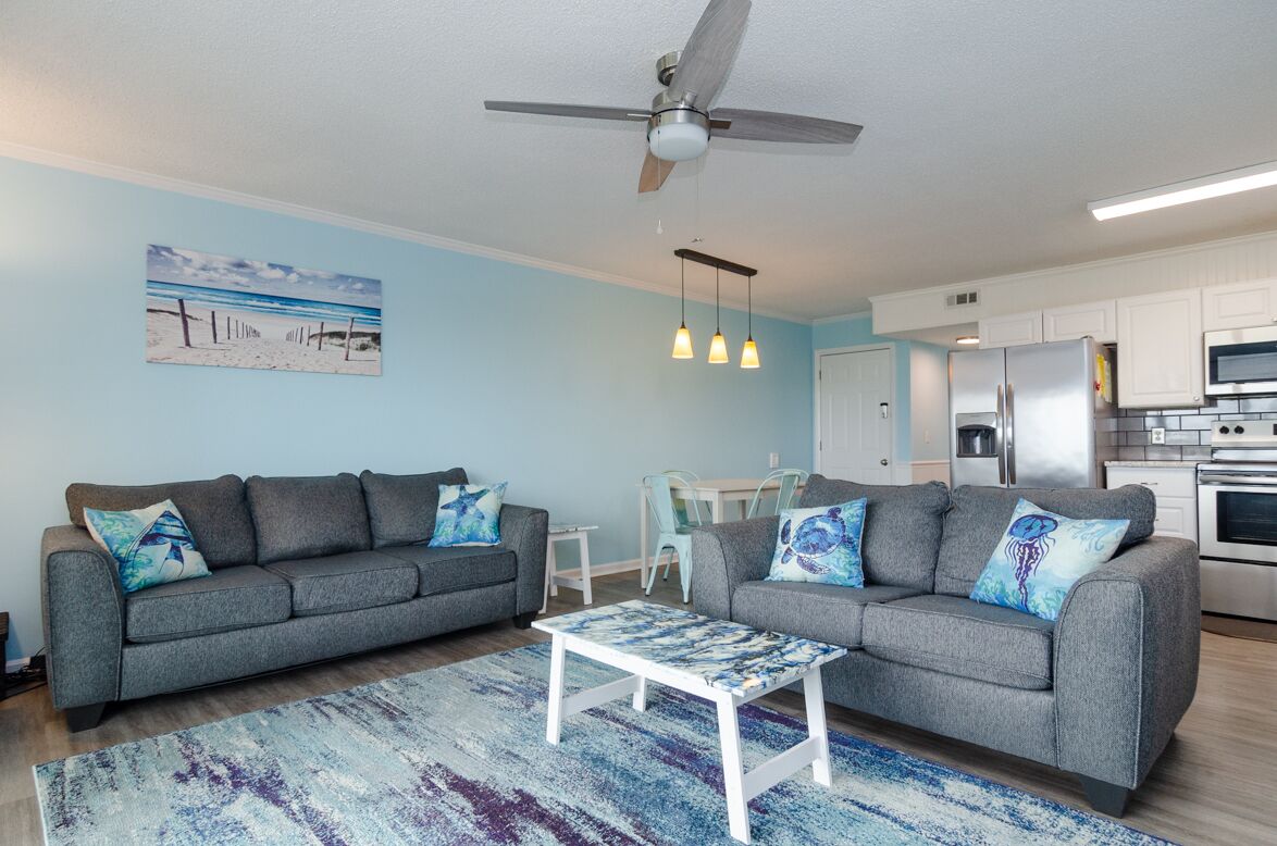 A modern living room with two grey sofas, a coffee table, a ceiling fan, pendant lights, a kitchen area with white cabinets, and a beach-themed wall art.