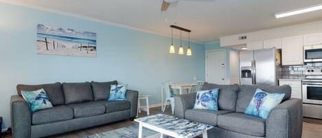 A modern living room with two grey sofas, a coffee table, a ceiling fan, pendant lights, a kitchen area with white cabinets, and a beach-themed wall art.