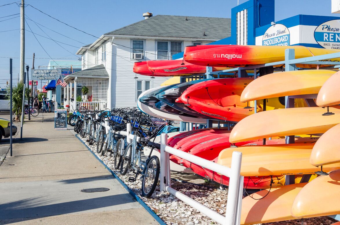 Kayaks y bicicletas están estacionadas a lo largo de una acera junto a un edificio con un letrero que dice "Pleasure Blend Paddleboard Rentals".