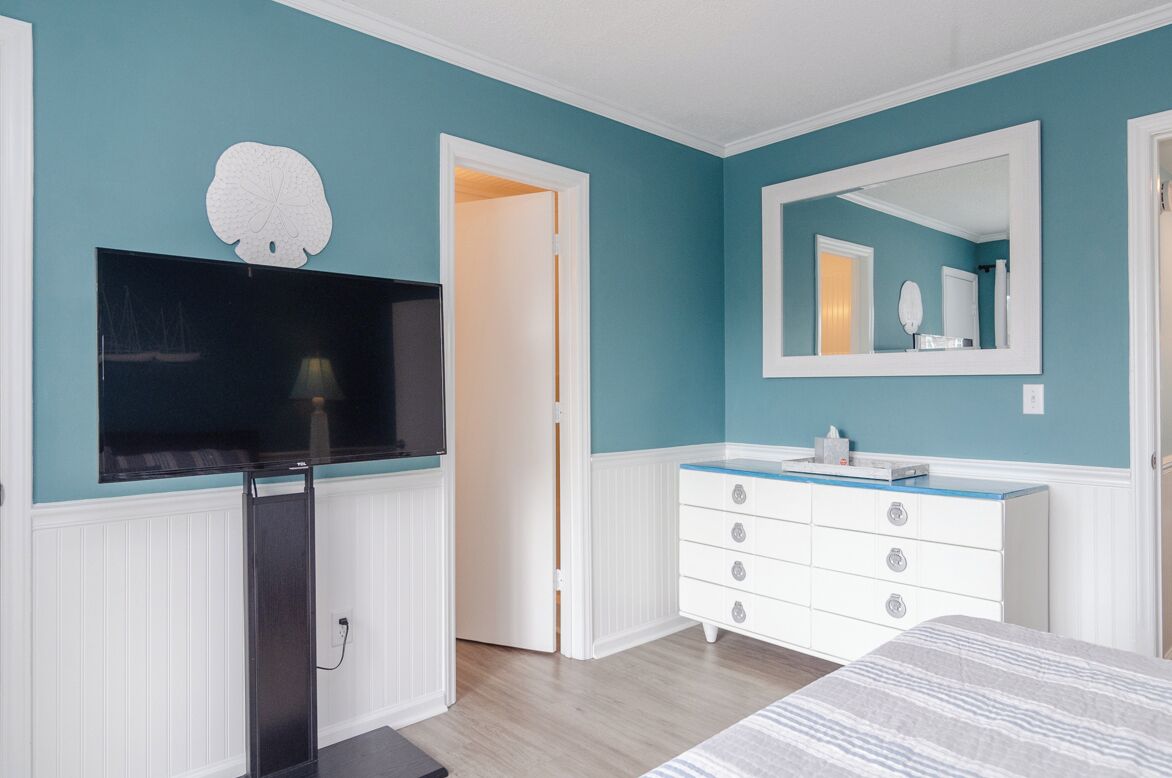 A bedroom with teal walls, a white dresser, a large mirror, and a flat-screen TV mounted on the wall.