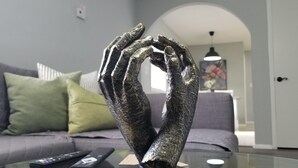 Una escultura de manos formando un corazón sobre una mesa de vidrio en una sala de estar con un sofá gris y controles remotos.
