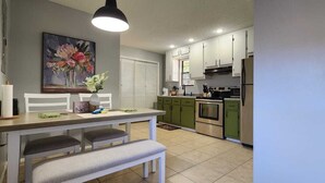 Una cocina con armarios blancos, una mesa de comedor con bancos, un cuadro en la pared y una lámpara colgante.