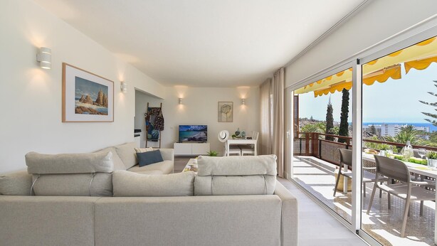 A modern living room with a sofa, a dining table, and a balcony with a view.