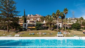 A swimming pool with clear blue water, surrounded by lounge chairs and umbrellas, with buildings and palm trees in the background.