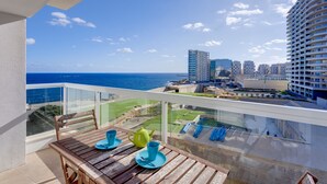 Un balcon offrant une vue sur l  ’océan et le paysage urbain, avec une table en bois, des tasses bleues et jaunes, et une balustrade en verre.