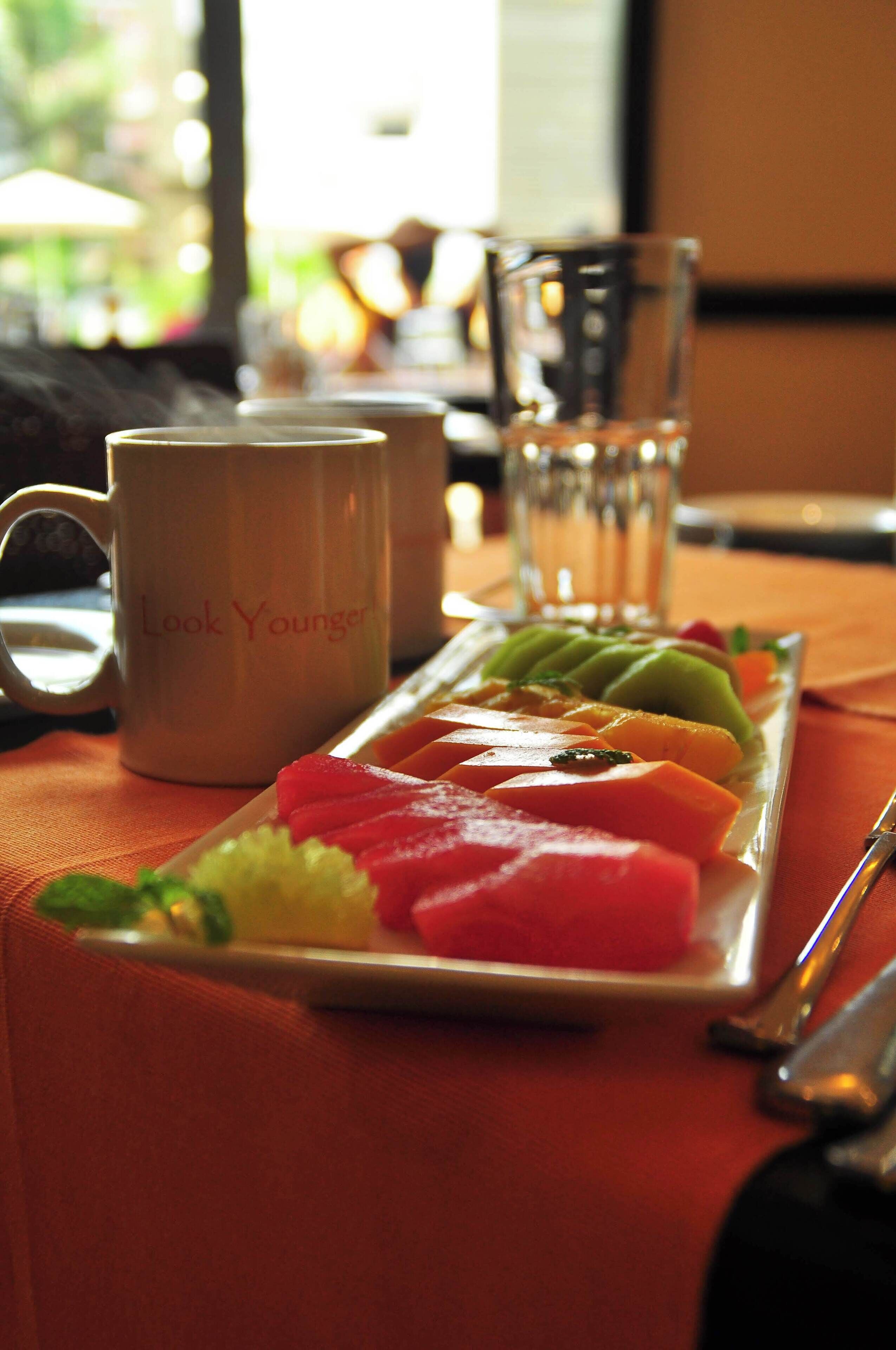 Una bandeja de desayuno con varias frutas y una taza con la etiqueta 'Look Younger' sobre una mesa.