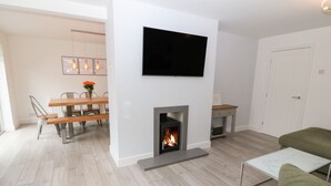 A modern living room with a fireplace, a flat-screen TV mounted on the wall, and a dining area with wooden furniture and a vase of flowers. - Daresfield (Cemaes Bay)