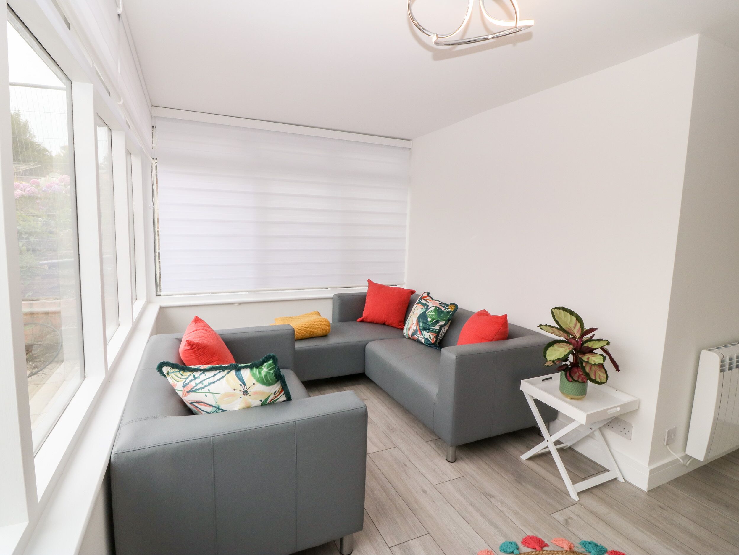 A modern living room with a grey sectional sofa, red and yellow cushions, a small white side table with a plant, and a large window with blinds.