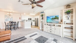 A modern living room with a ceiling fan, a TV, a wooden entertainment unit, and a kitchen in the background.