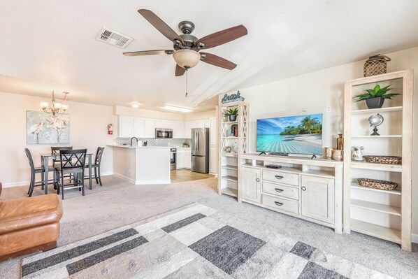 A modern living room with a ceiling fan, a TV, a wooden entertainment unit, and a kitchen in the background. - Waterfront Escape | Pool & Fishing Dock Near Beach (6 night minimum) (Corpus Christi)