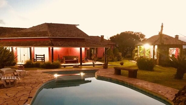 A swimming pool with a stone deck, a covered patio with chairs, and a small gazebo in a garden setting. - Pousada Por do Sol (Camanducaia)