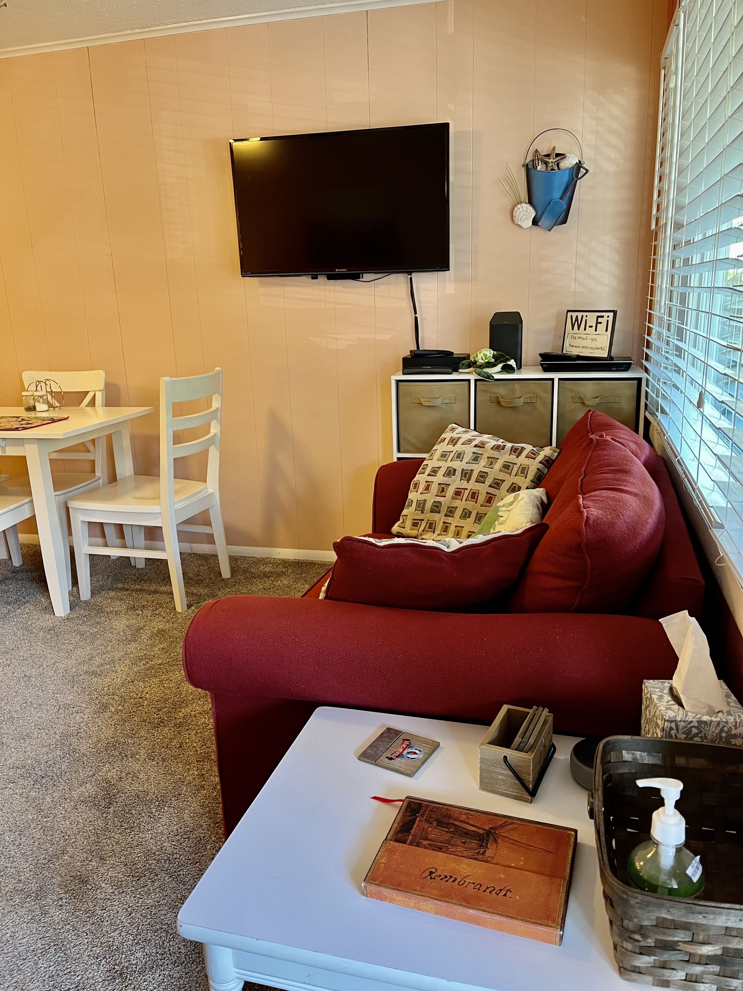 Un soggiorno con una TV a schermo piatto montata a parete, un divano rosso con cuscini decorati, un tavolino da caffè bianco con un libro e una bottiglia di gel disinfettante, e un piccolo tavolino laterale con un simbolo Wi-Fi e oggetti decorativi.