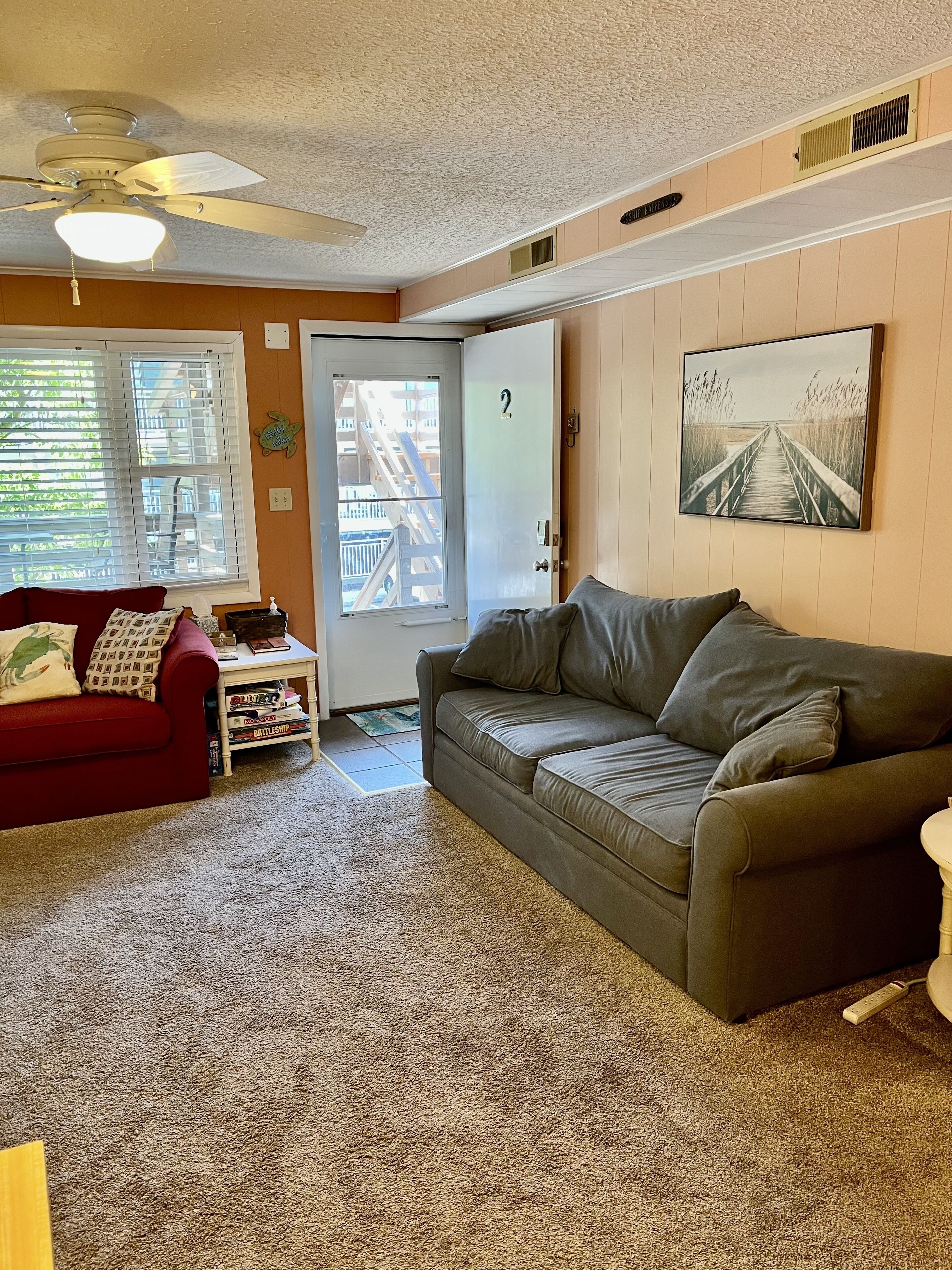 A living room with a red couch, a grey couch, a ceiling fan, a door with a number, and a painting of a bridge.