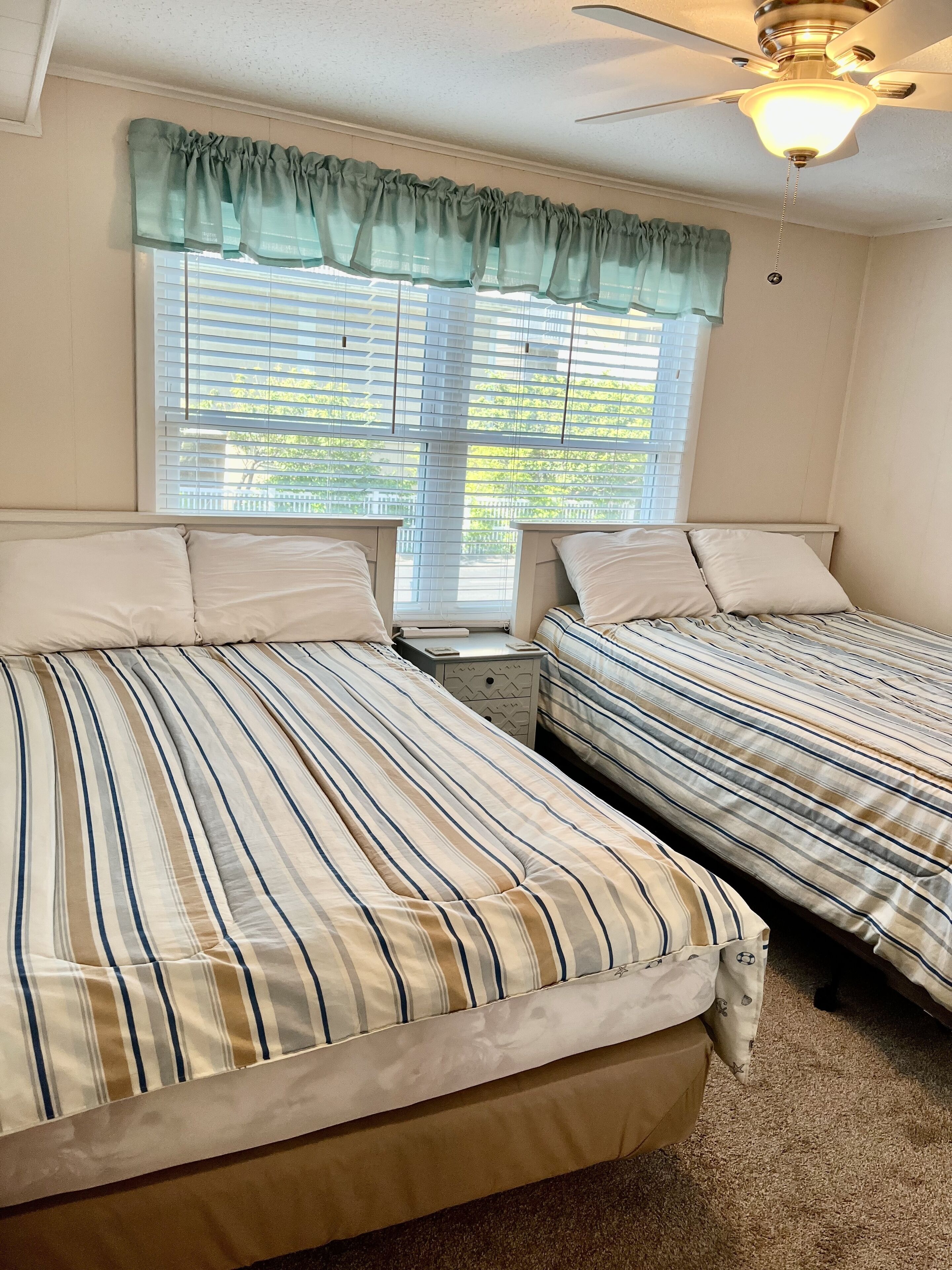 A bedroom with two beds, a ceiling fan, a window with blinds, and a nightstand.