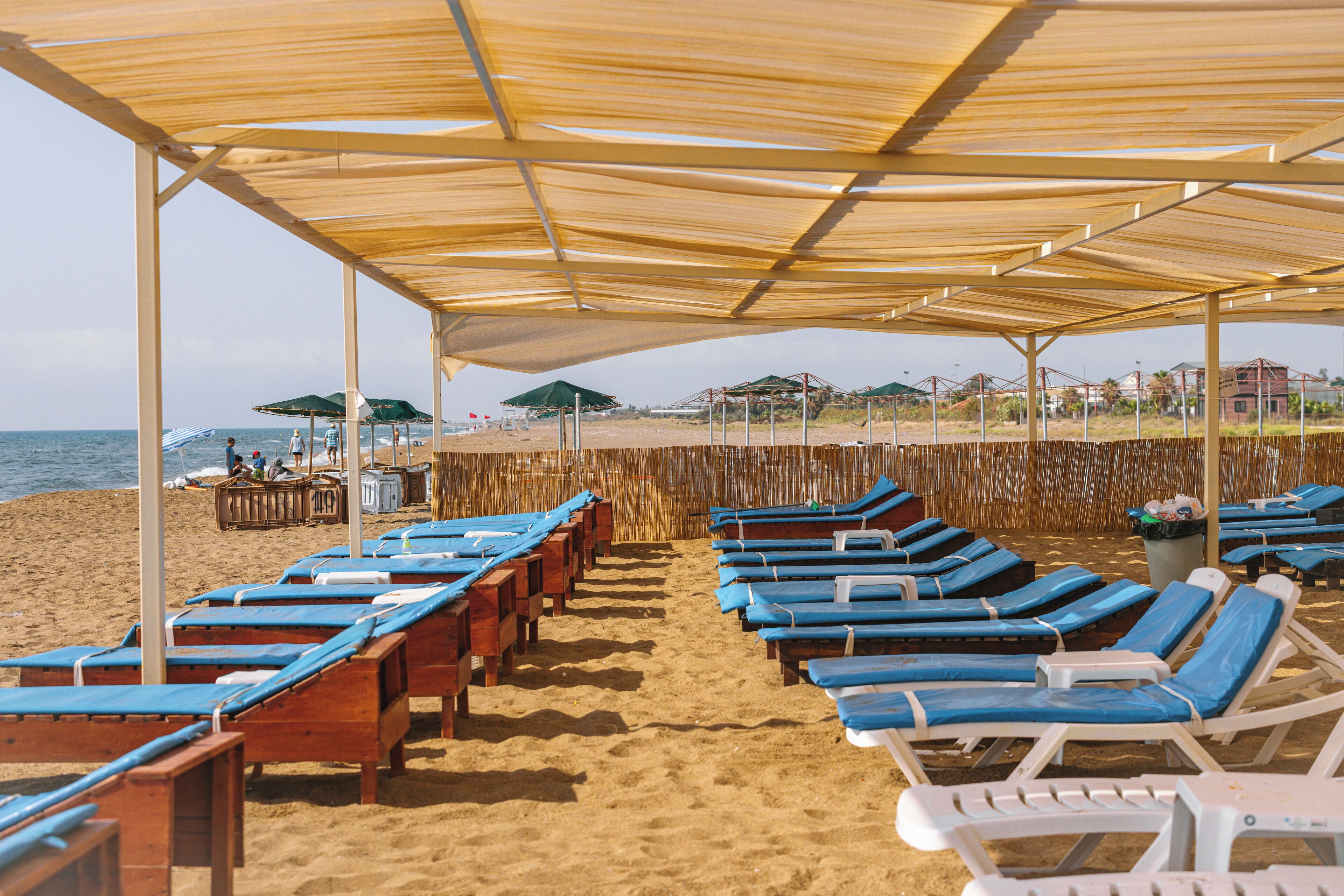 A beachside lounge area with rows of sunbeds under a shaded canopy, a sandy beach, and umbrellas in the background.