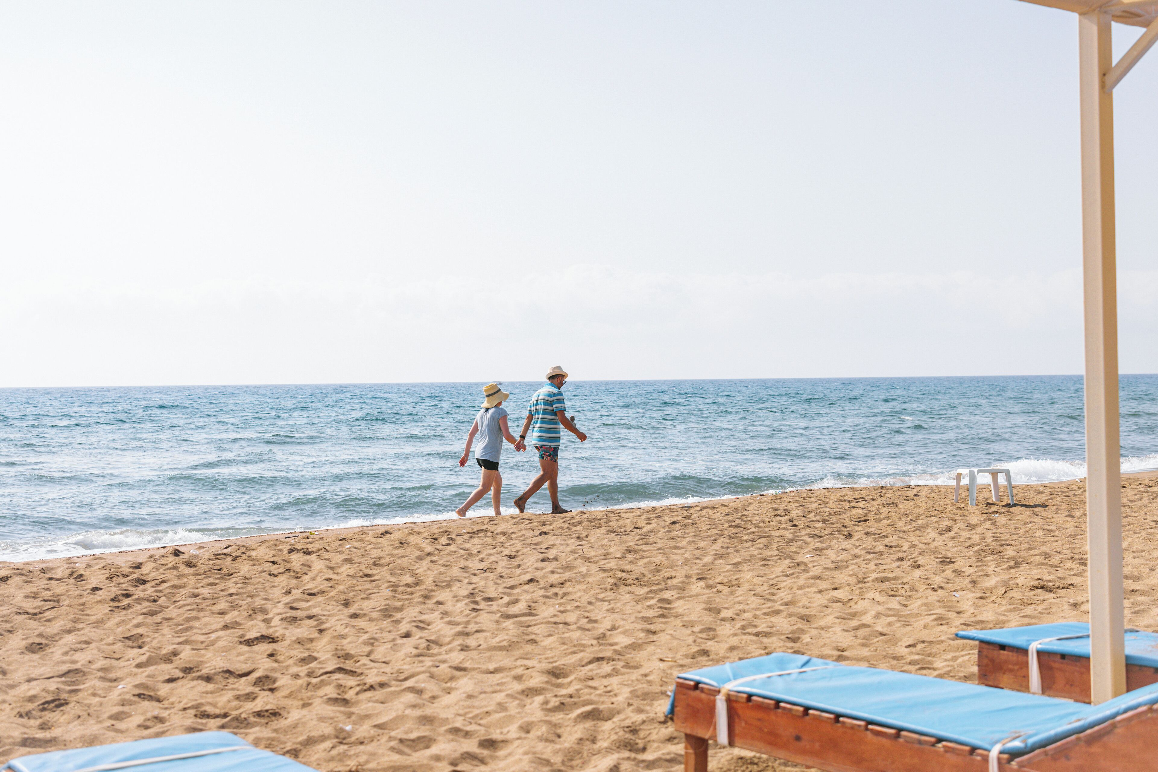 Una playa con dos personas caminando, una estructura de madera con sillas azules y una vista despejada del mar.