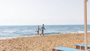 Una playa con dos personas caminando, una estructura de madera con sillas azules y una vista despejada del mar.