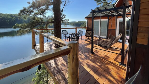 A wooden deck with outdoor furniture overlooking a lake, a covered seating area, and a building with a glass door.