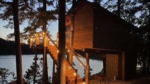 A wooden cabin with a staircase leading to a deck, surrounded by trees and a body of water.