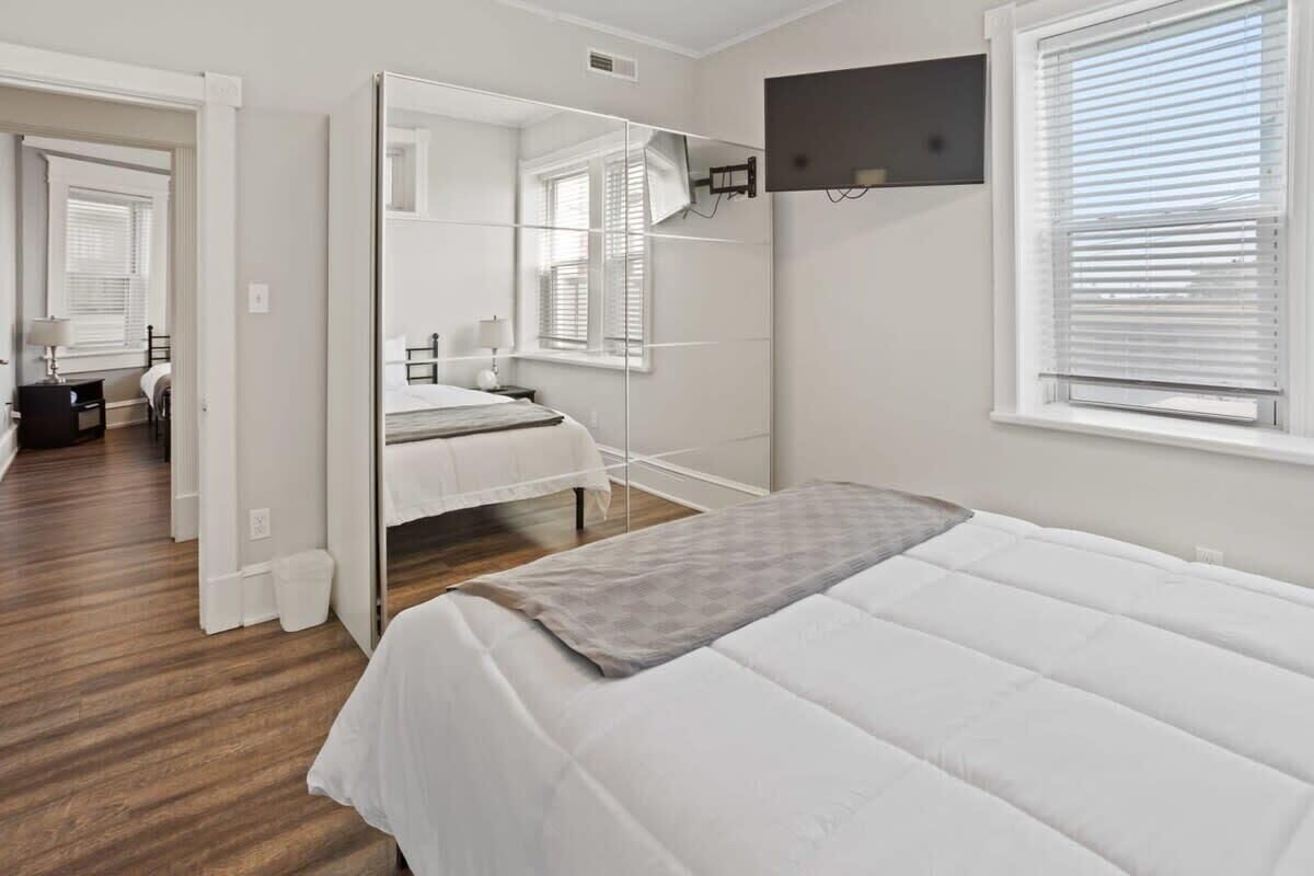 Une chambre à coucher comprenant un lit, un miroir, une fenêtre avec des stores et une porte donnant sur une autre pièce.