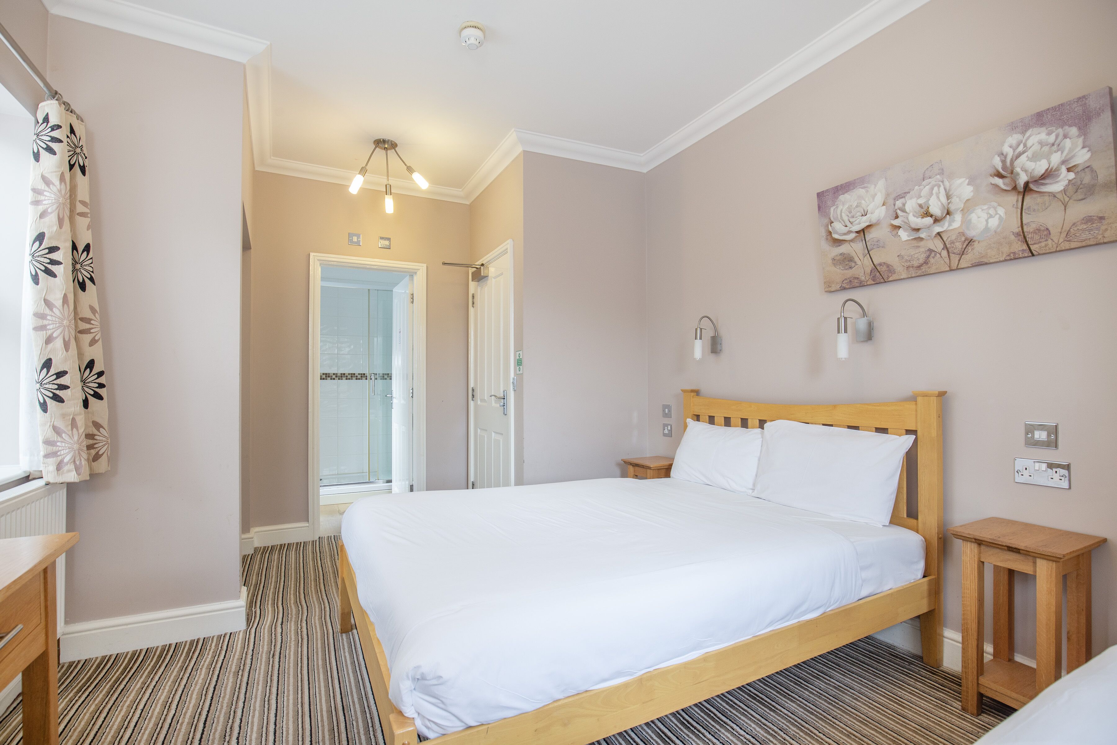 A bedroom with a wooden bed, a nightstand, a small wooden table, a painting on the wall, and a door leading to another room.