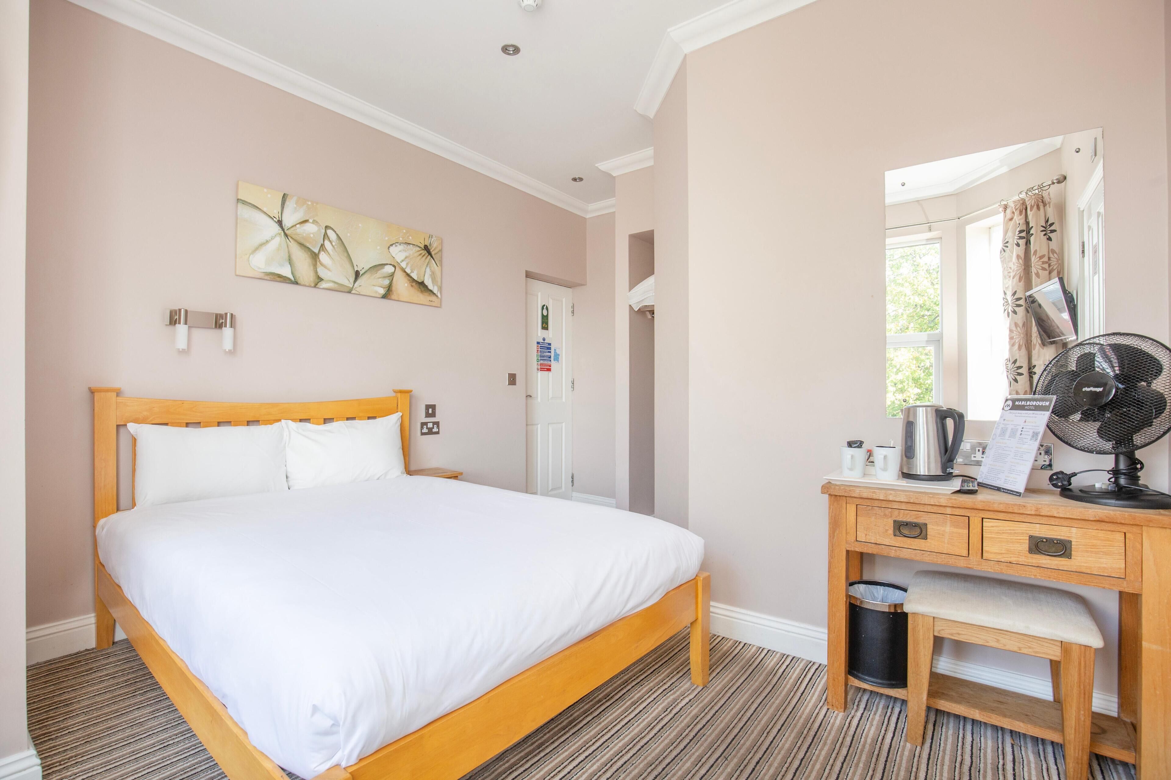 Une chambre d’hôtel bien rangée, avec un lit, un bureau en bois, un ventilateur et une fenêtre avec des rideaux.