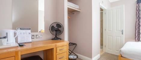 A hotel room with a wooden desk, a black fan, a white bed, a door, and a trash bin.