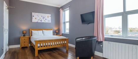A bedroom with a wooden bed, a nightstand, a black leather chair, a TV, and a window with curtains.