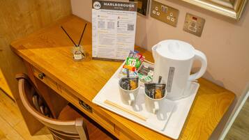 Une chambre d’hôtel avec un bureau en bois, une tasse blanche, un plateau avec des portions de nourriture emballées, une bouteille d’eau et un panneau indicateur.