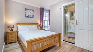 A bedroom with a wooden bed, bedside tables, a nightstand, a window with curtains, a bathroom with a mirror and sink, and a door. - Marlborough Hotel (Norwich)
