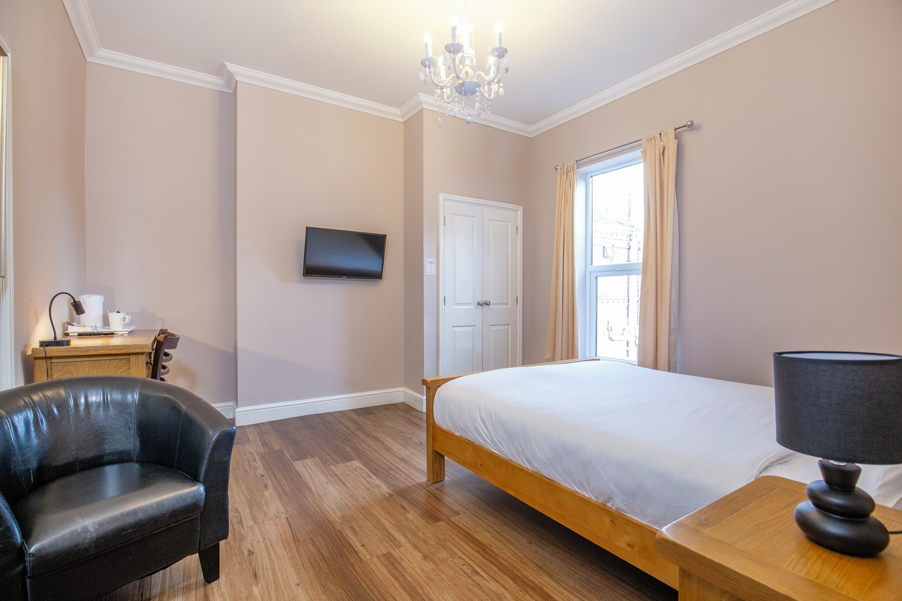 A bedroom with a bed, a chair, a bedside table with a lamp, a television on the wall, and a window with curtains.