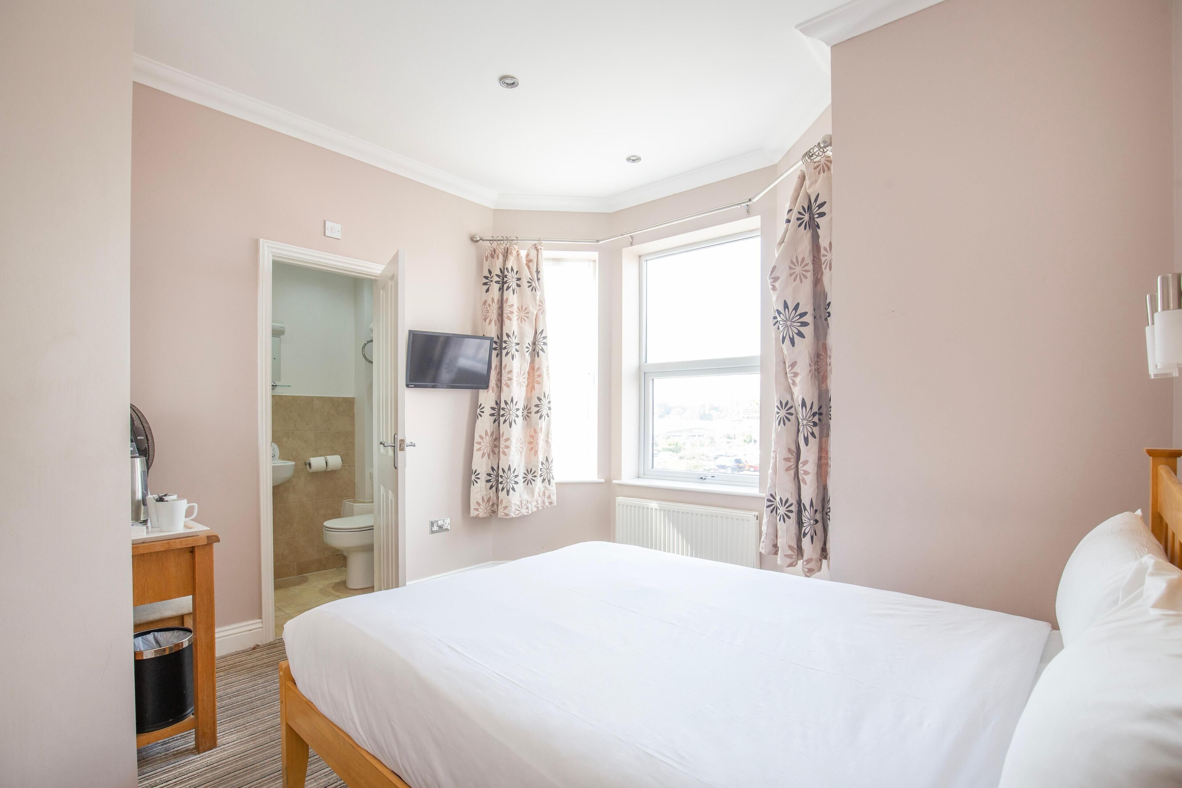 A bedroom with a bed, a television, a bathroom with a toilet, and a window with curtains.