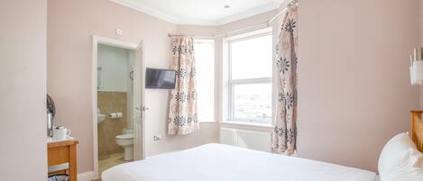 A bedroom with a bed, a television, a bathroom with a toilet, and a window with curtains.