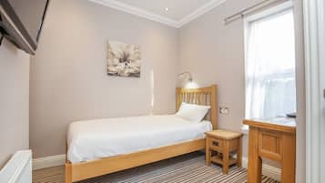 A bedroom with a single bed, a wooden nightstand, a desk, a lamp, and a large window with curtains.