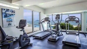 A gym with treadmills, an elliptical, and a stationary bike, with large windows offering a view of the sea.