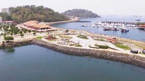 A coastal marina with boats, a park area, and buildings on a hillside.