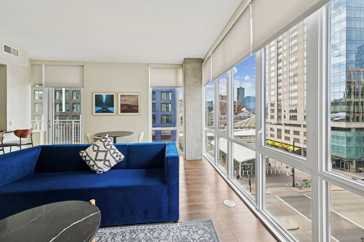 A modern living room with a blue sofa, a black coffee table, and a large window offering a city view.