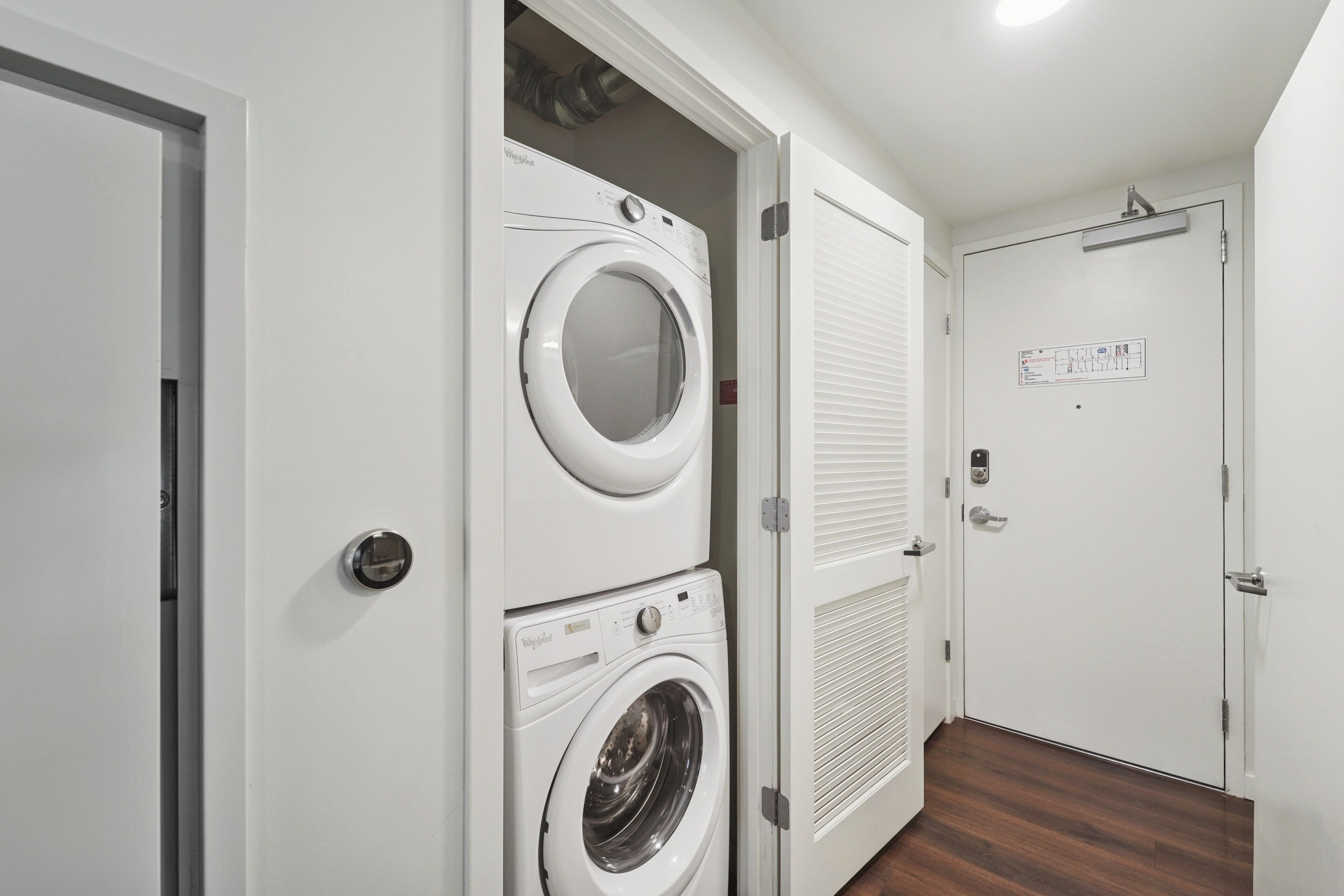 A laundry room with two stacked front-loading washing machines, a closed door with a control panel, and a wooden floor.