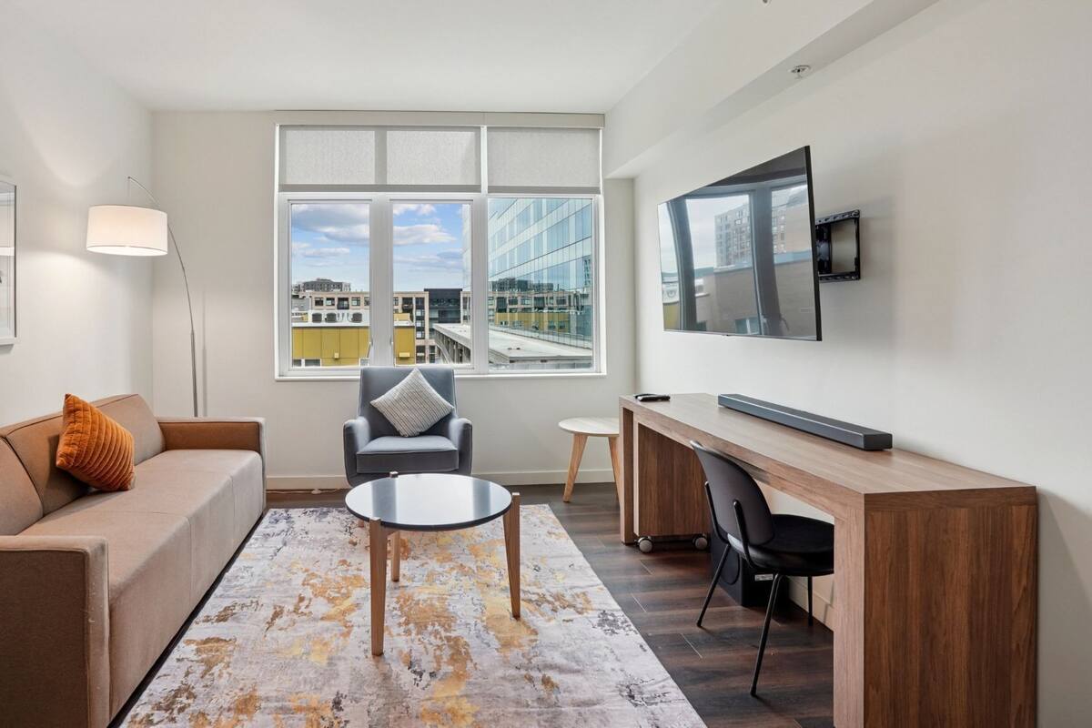 A modern living room with a sofa, armchair, coffee table, and a desk with a flat-screen TV. Large windows offer a view of the cityscape.