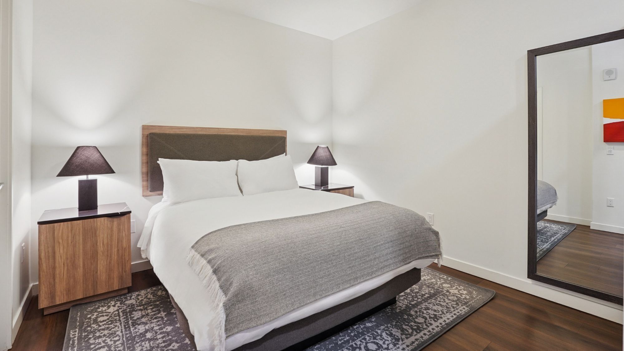 A neatly arranged hotel room with a bed, two bedside tables with lamps, a large mirror, and a patterned rug.