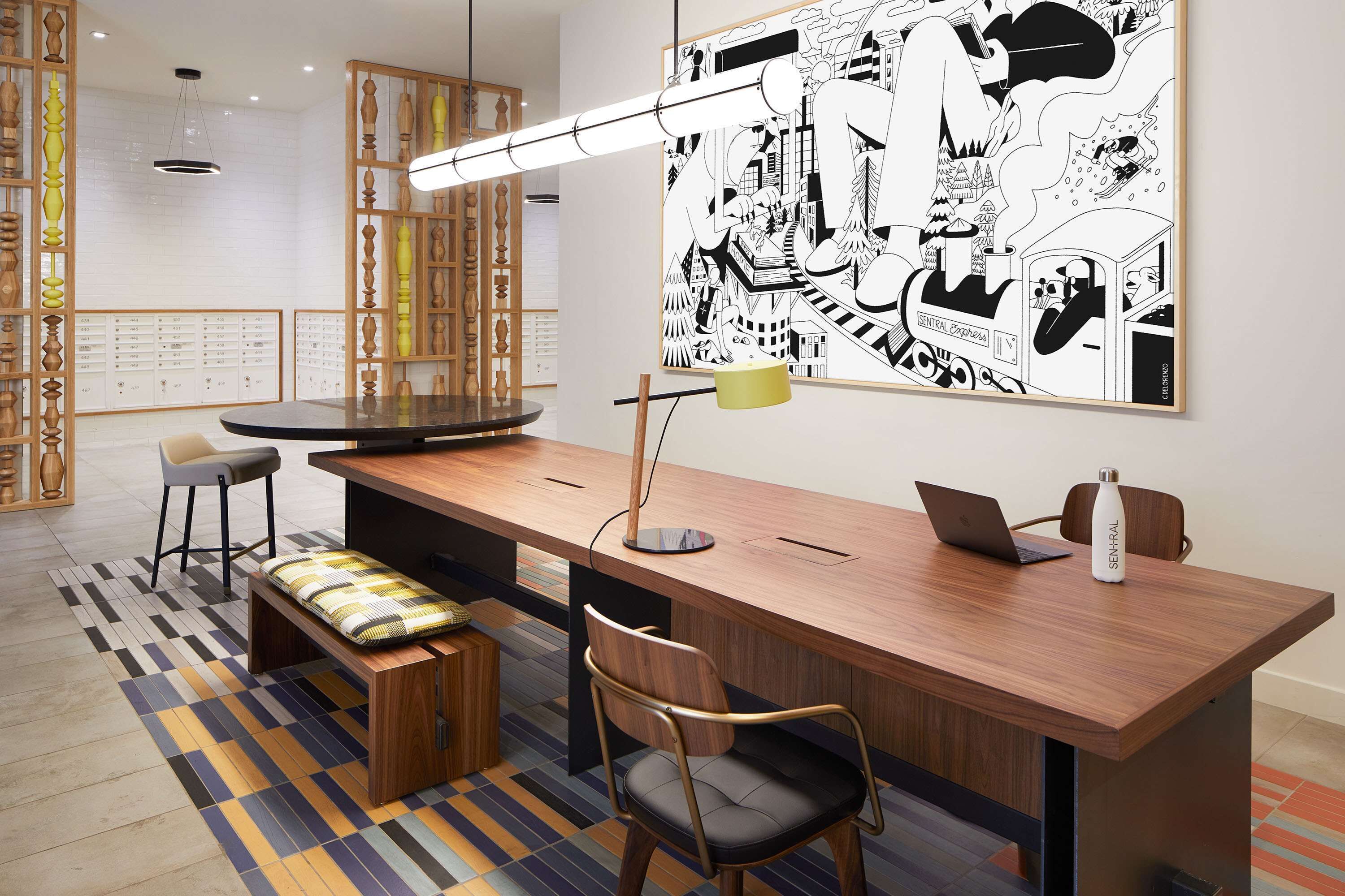 A modern office space with a long wooden desk, a high chair, a bench, a large abstract painting, and a pendant light.