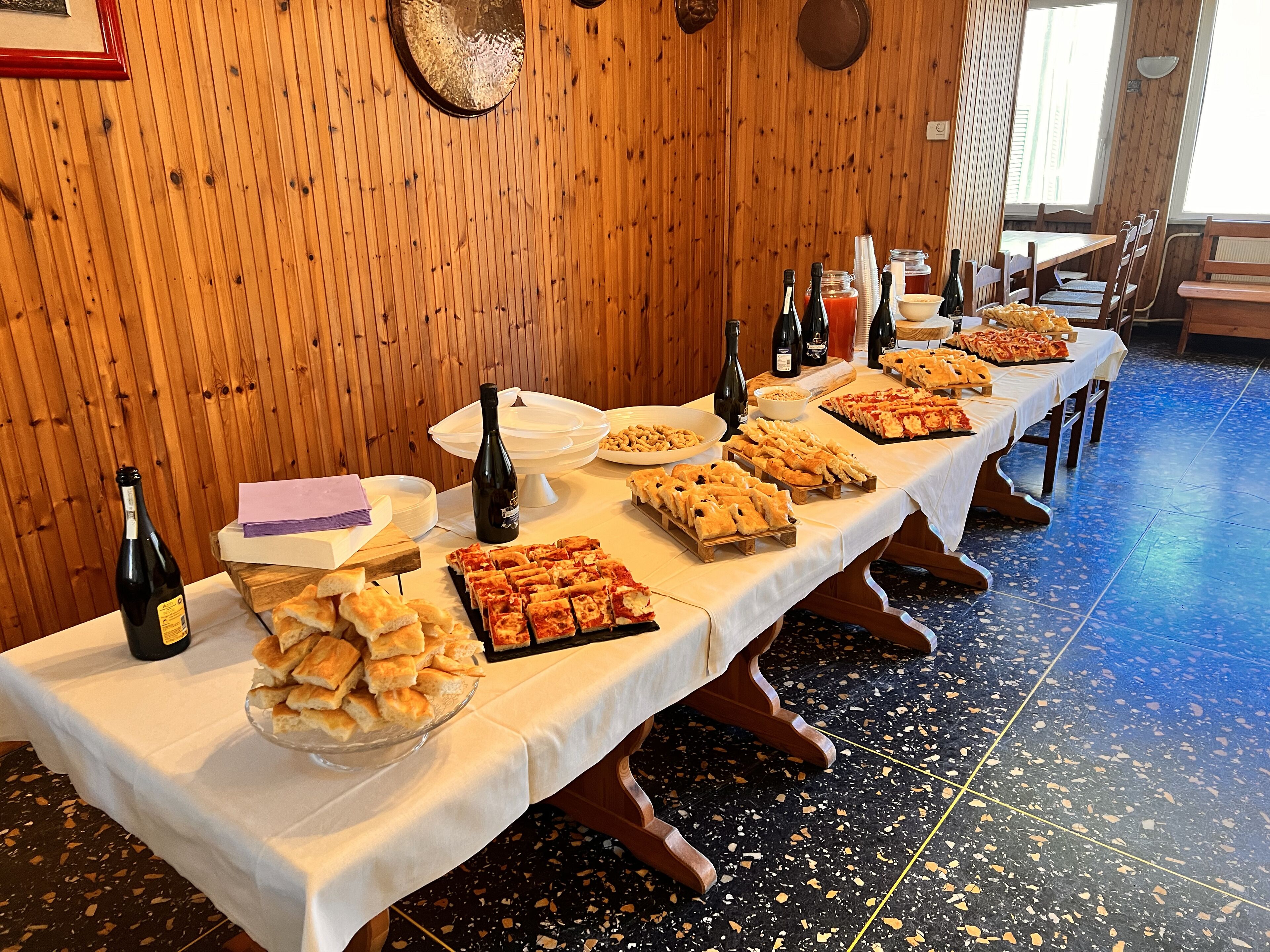 Una larga mesa con diversos alimentos y botellas de vino, frente a una pared de madera y un suelo de baldosas.
